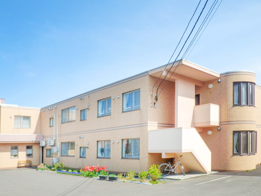 介護施設の建物外観。明るいレンガ造りの2階建て施設で、玄関前に花壇と自転車が見える。