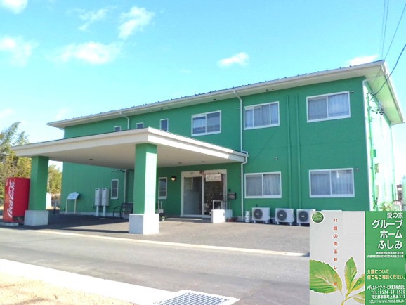 緑色の2階建て介護施設の外観。駐車スペースと車寄せが見える。