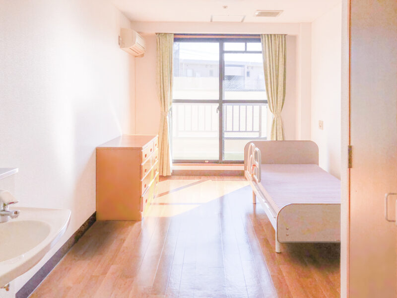介護施設の居室。ベッド、収納、洗面台、バルコニーを備えた明るい個室