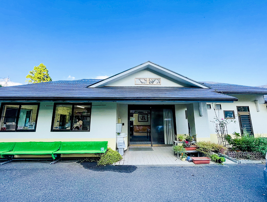 白壁の介護施設。緑のベンチと駐車場が見える平屋建物の外観。