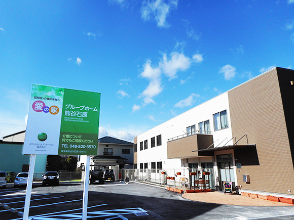 介護施設の建物外観。茶色レンガ造りの2階建て建物と駐車場、施設の看板が見える。