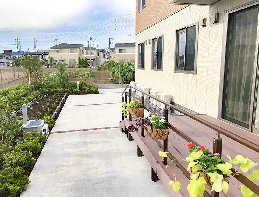 介護施設のテラスと庭園。プランター花壇と周辺の住宅地が見える。