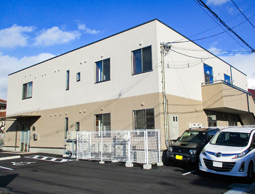 白とベージュのツートンカラーの介護施設外観。駐車場と入口が見える。