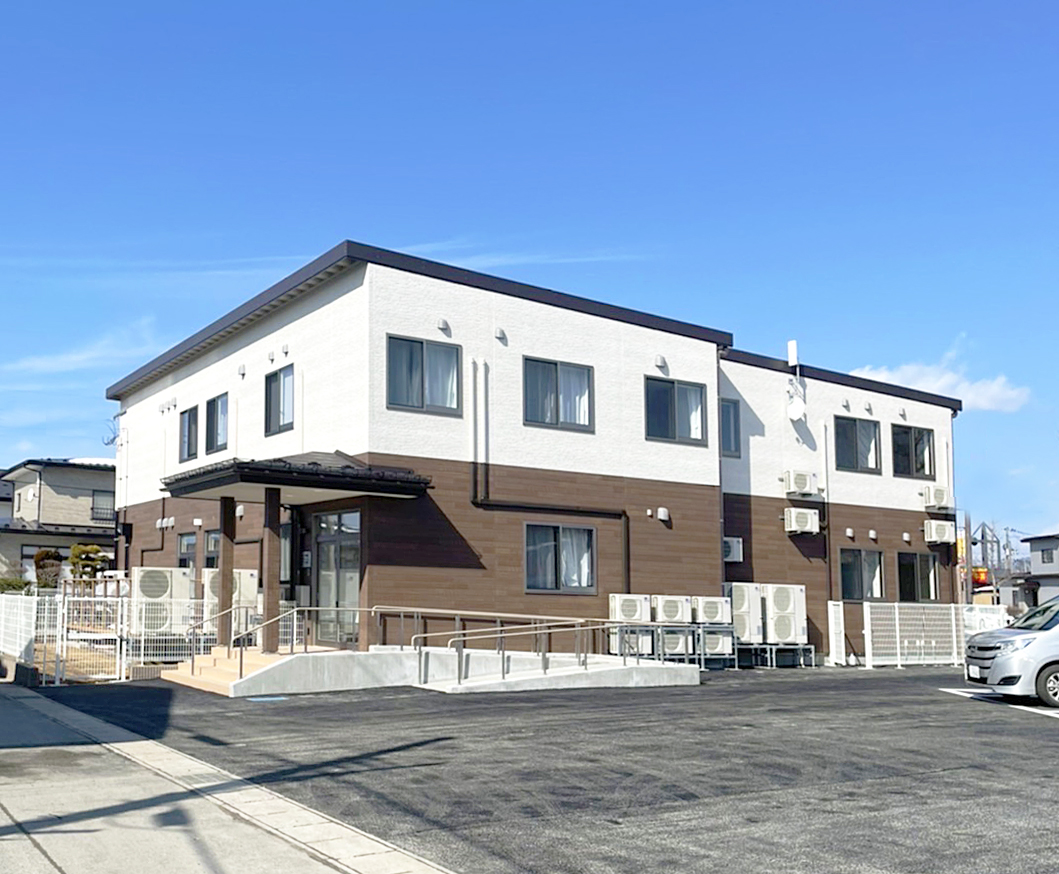白と茶色のツートンカラーの介護施設外観。晴天で駐車場から撮影。